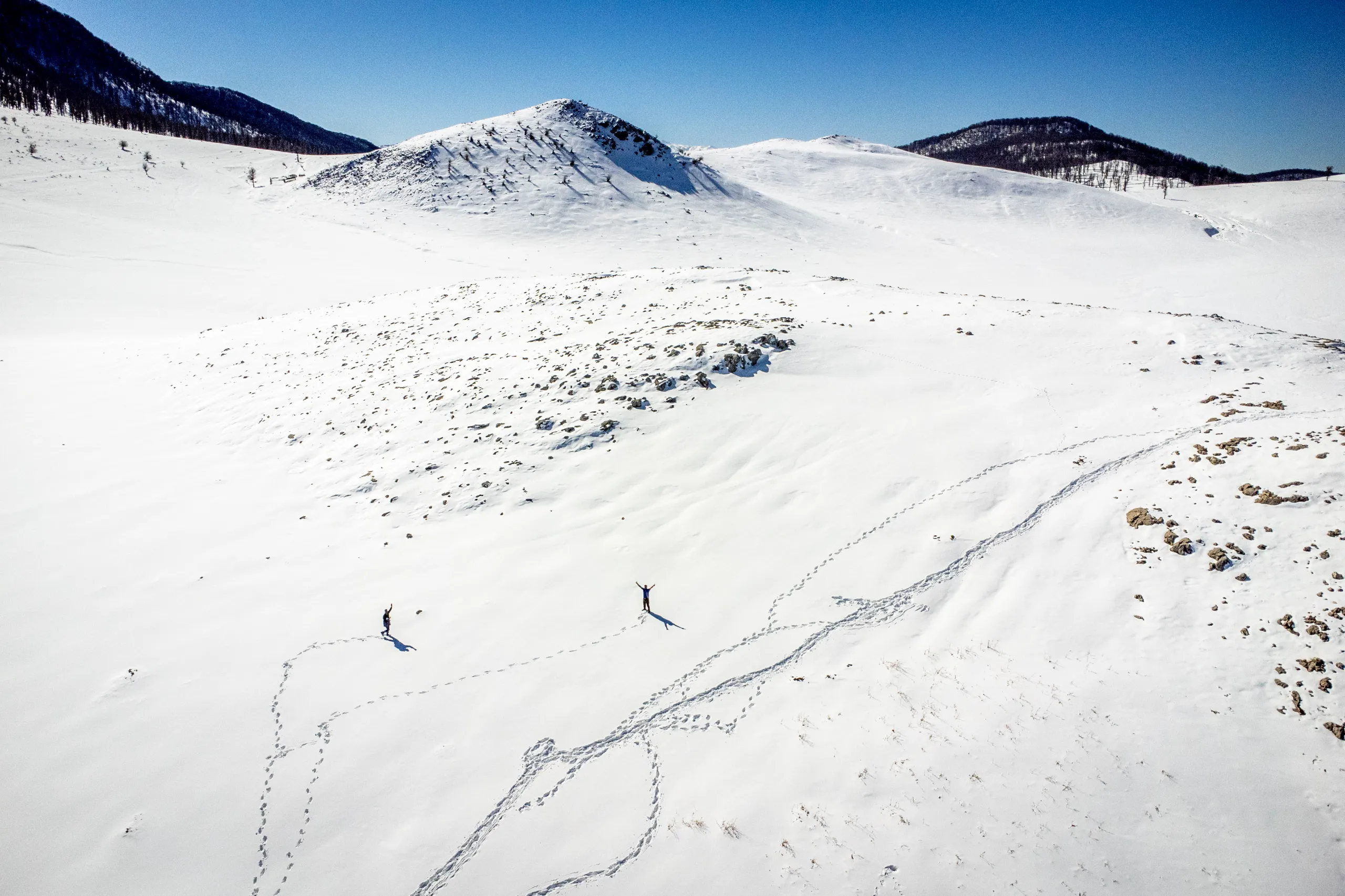 An aerial view of two hikers standing with raised arms on a vast snow-covered mountain plain with visible footsteps, under a clear blue sky. Photograph by Mohammad Ataei.