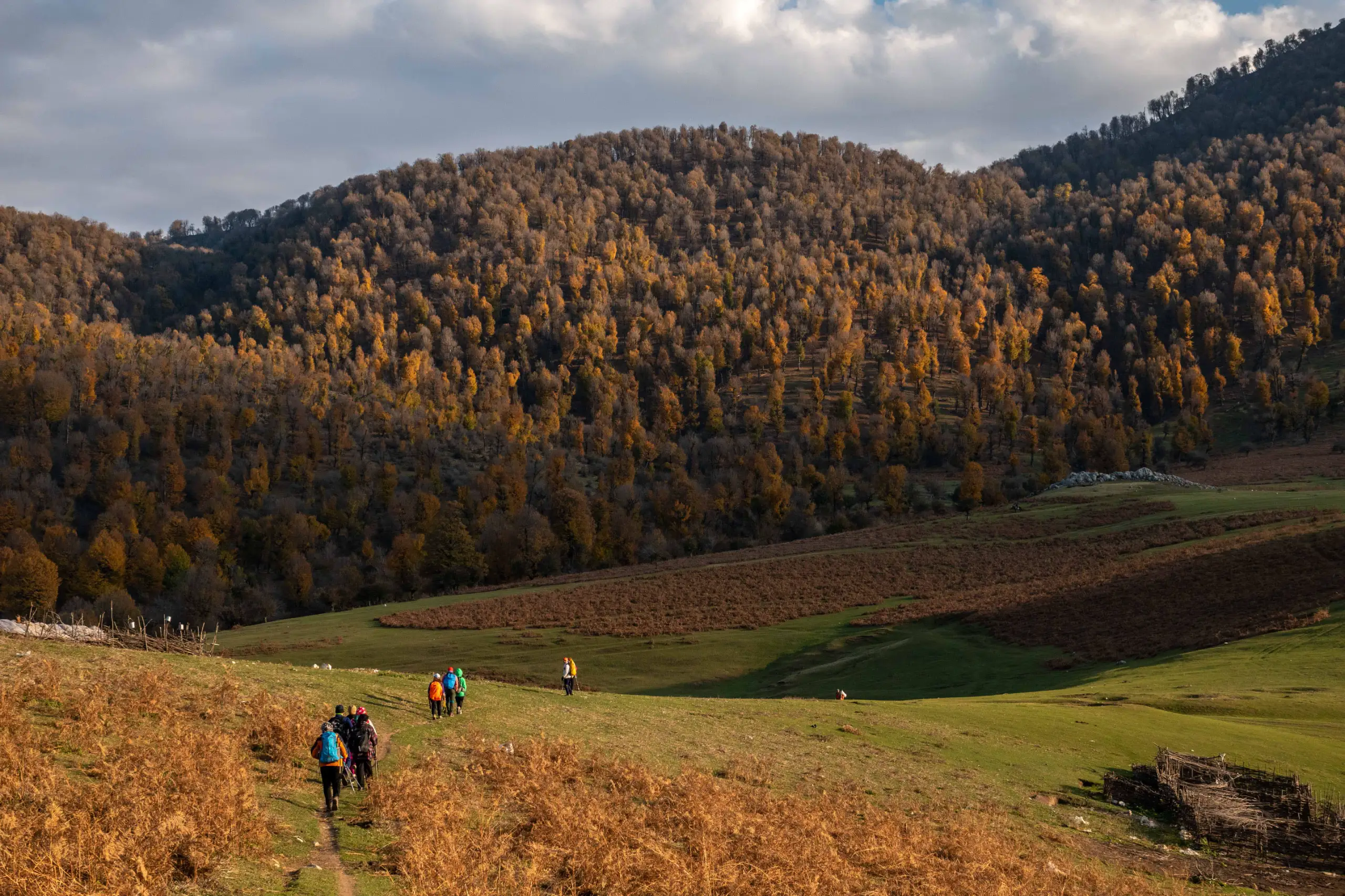 A group of hikers walking along a trail in the rolling green and autumn-colored hills of Markouh Plain under a cloudy sky. Photograph by Mohammad Ataei.