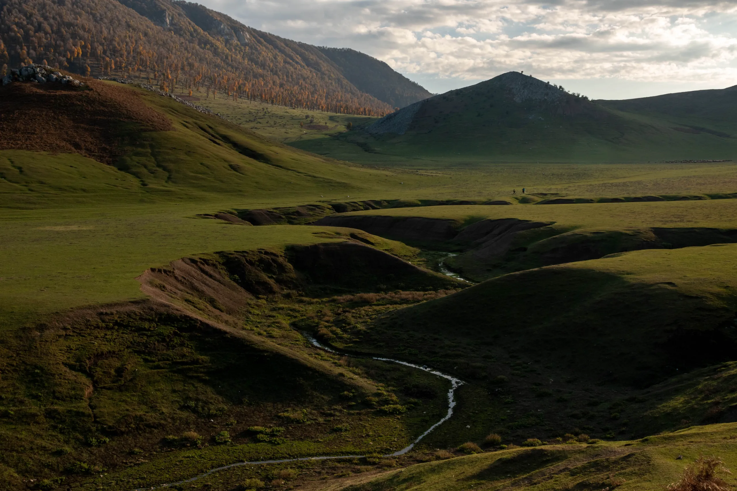 Wide landscape shot of Markouh Plain in Golestan Province featuring rolling green hills, a winding stream in the valley, and distant mountains under a cloudy sky. Photograph by Mohammad Ataei.