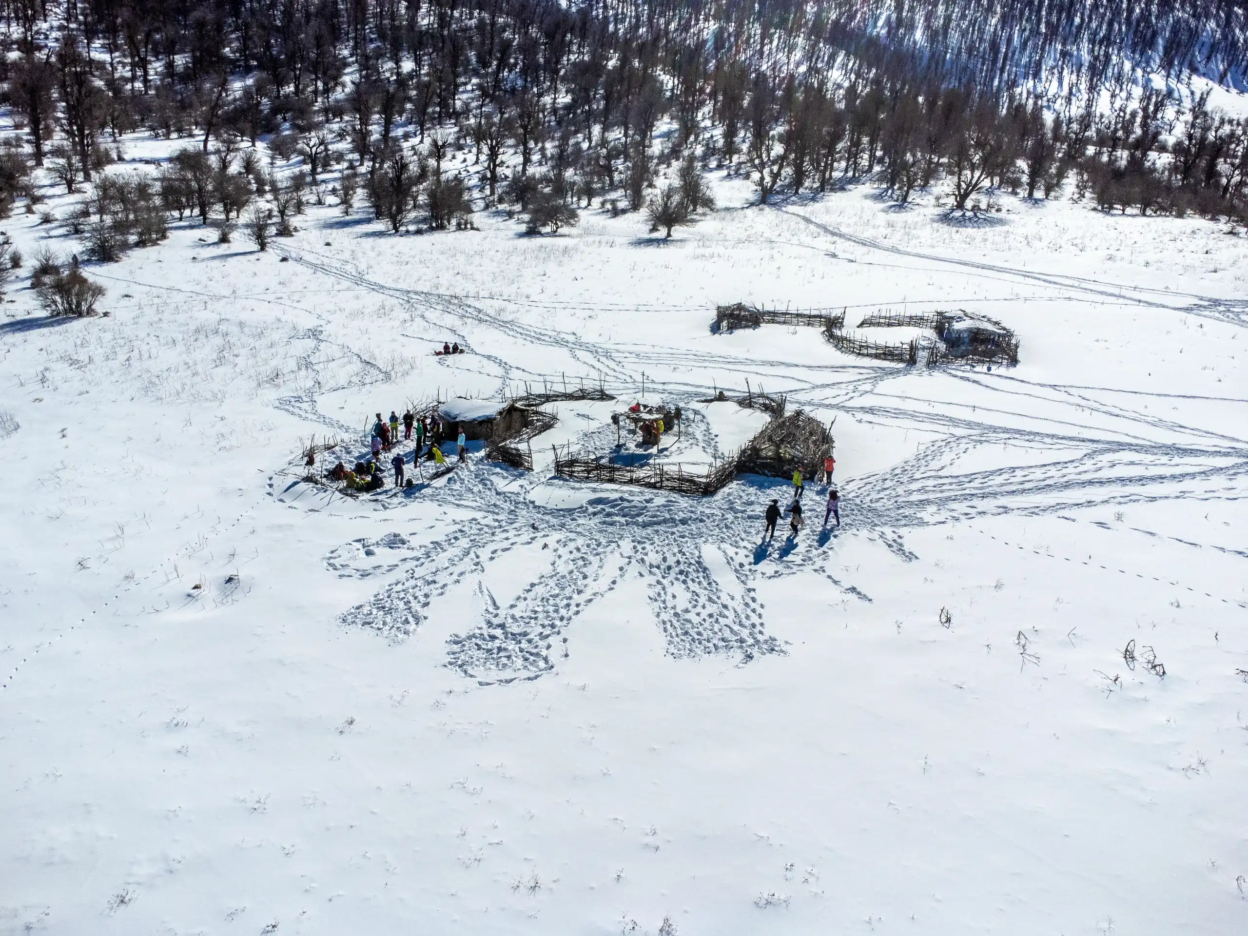 An aerial shot shows a snowy mountain plain with rustic wooden enclosures, scattered people, and intricate footprints in the snow, set against a forest backdrop in winter. Photograph by Mohammad Ataei.