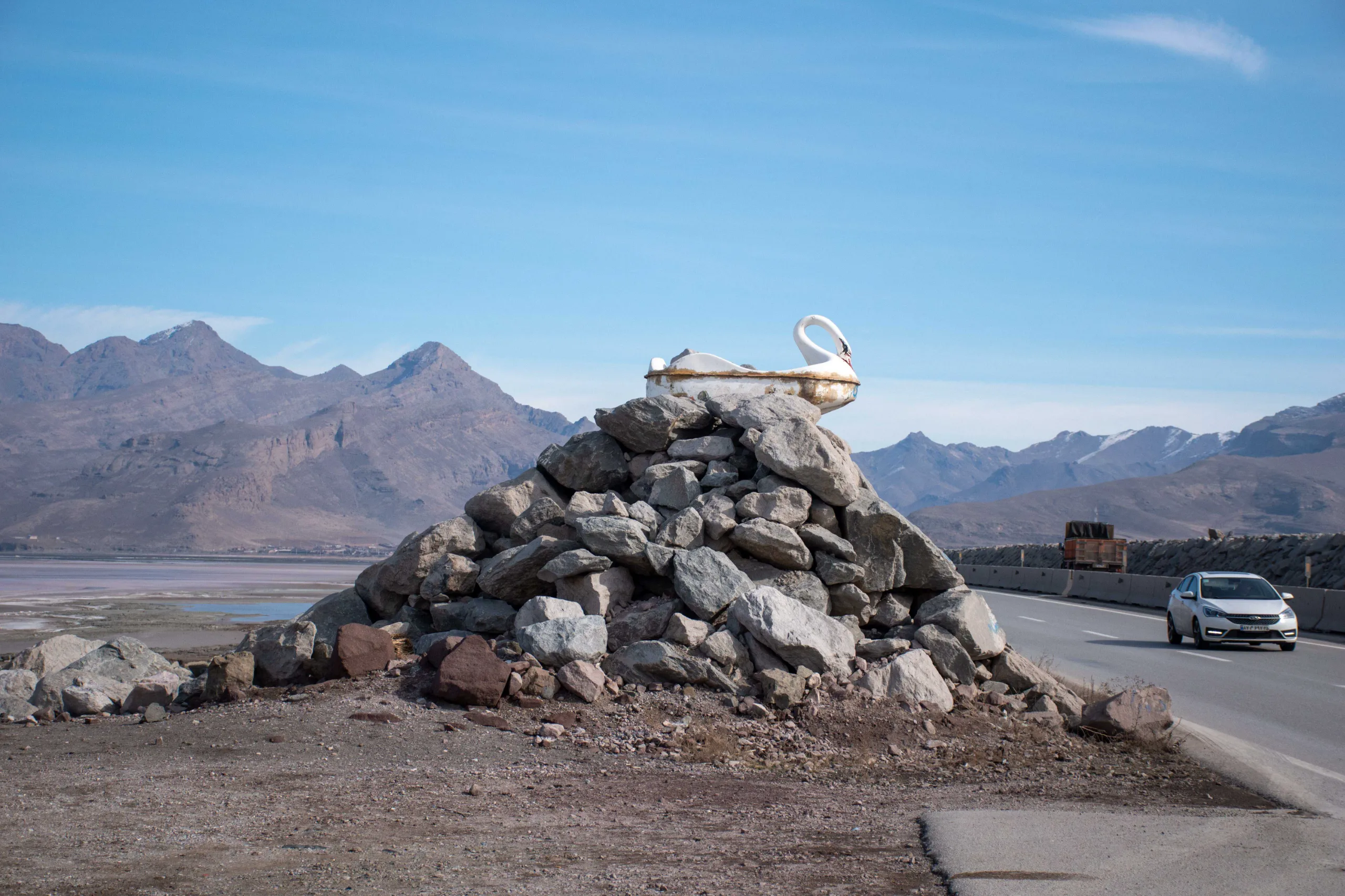A weathered white swan-shaped pedal boat resting on a pile of rocks beside a coastal highway, with barren mountains and a glimpse of Lake Urmia in the background under a blue sky. Photograph by Mohammad Ataei.