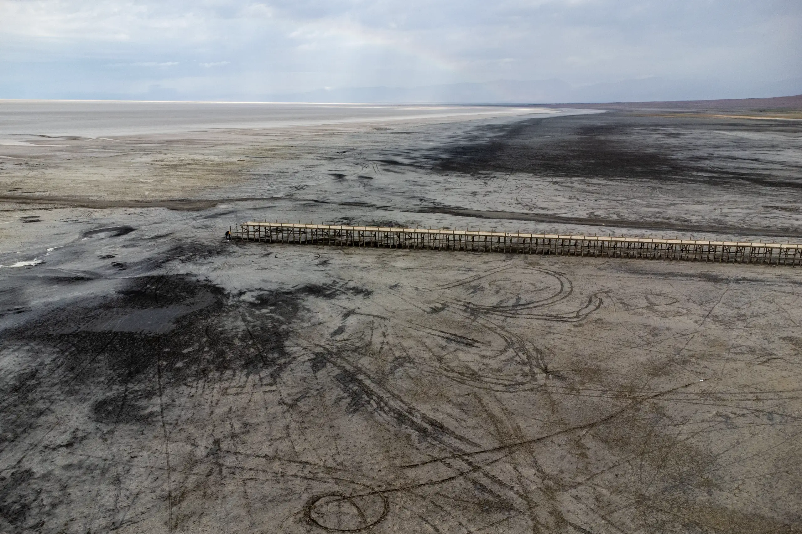 An aerial shot showing the vast, dry mud flats and receding shoreline of Lake Urmia, with a long wooden pier extending over the cracked ground under an overcast sky. Photograph by Mohammad Ataei.