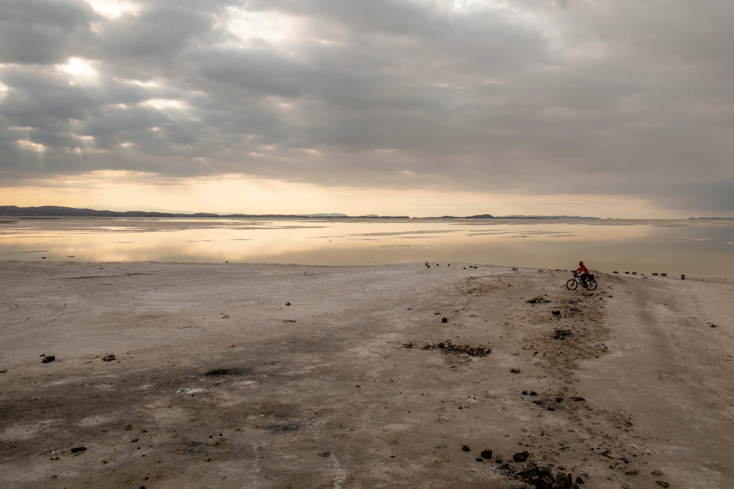 A wide-angle landscape shot shows a lone cyclist on a sandy and saline shore next to a reflective lake under a cloudy sky. Photograph by Mohammad Ataei.