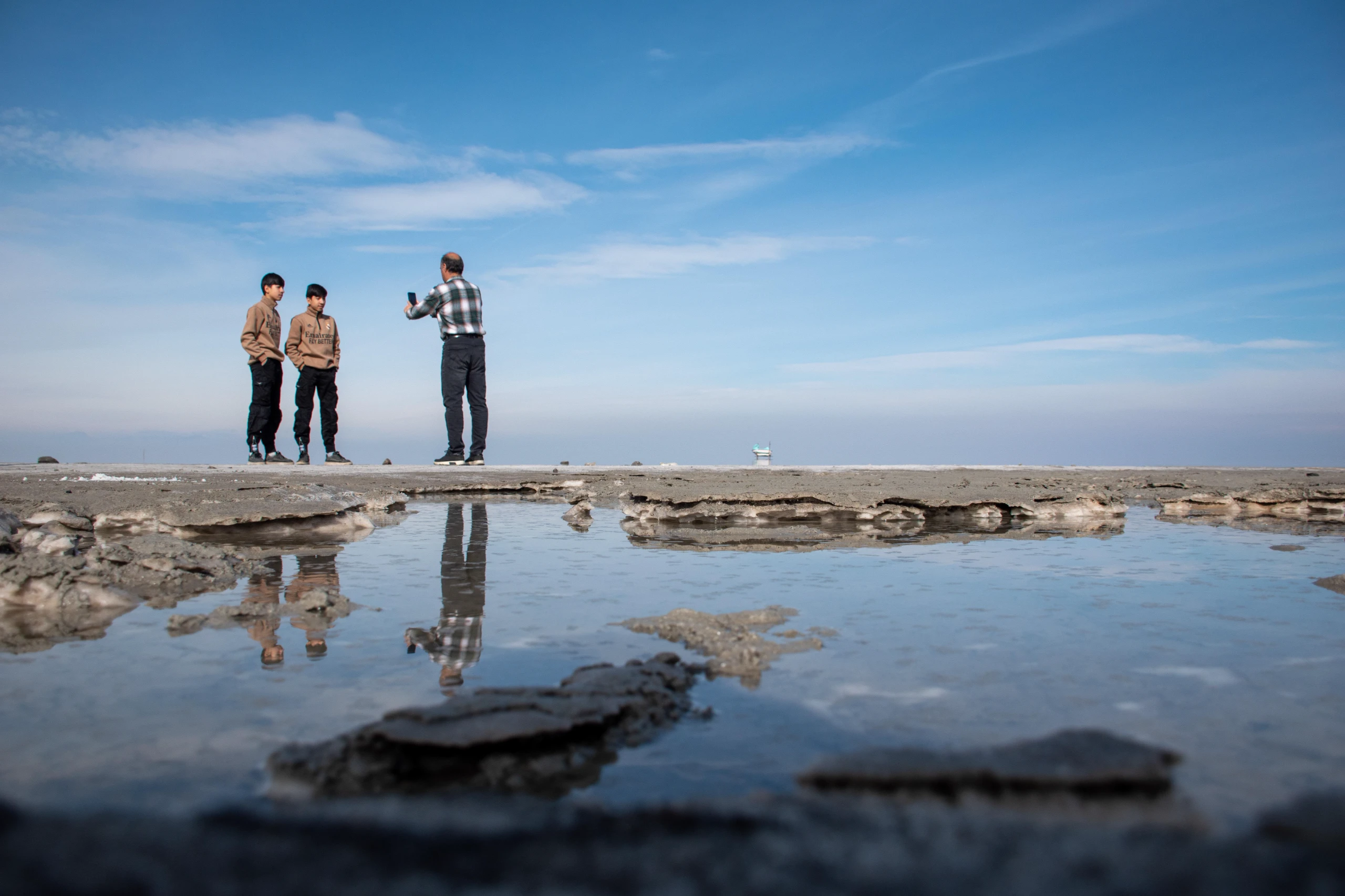 A man taking a photo of two youths with a smartphone standing on the muddy shore of Lake Urmia under a blue sky, with reflections in a puddle. Photograph by Mohammad Ataei.