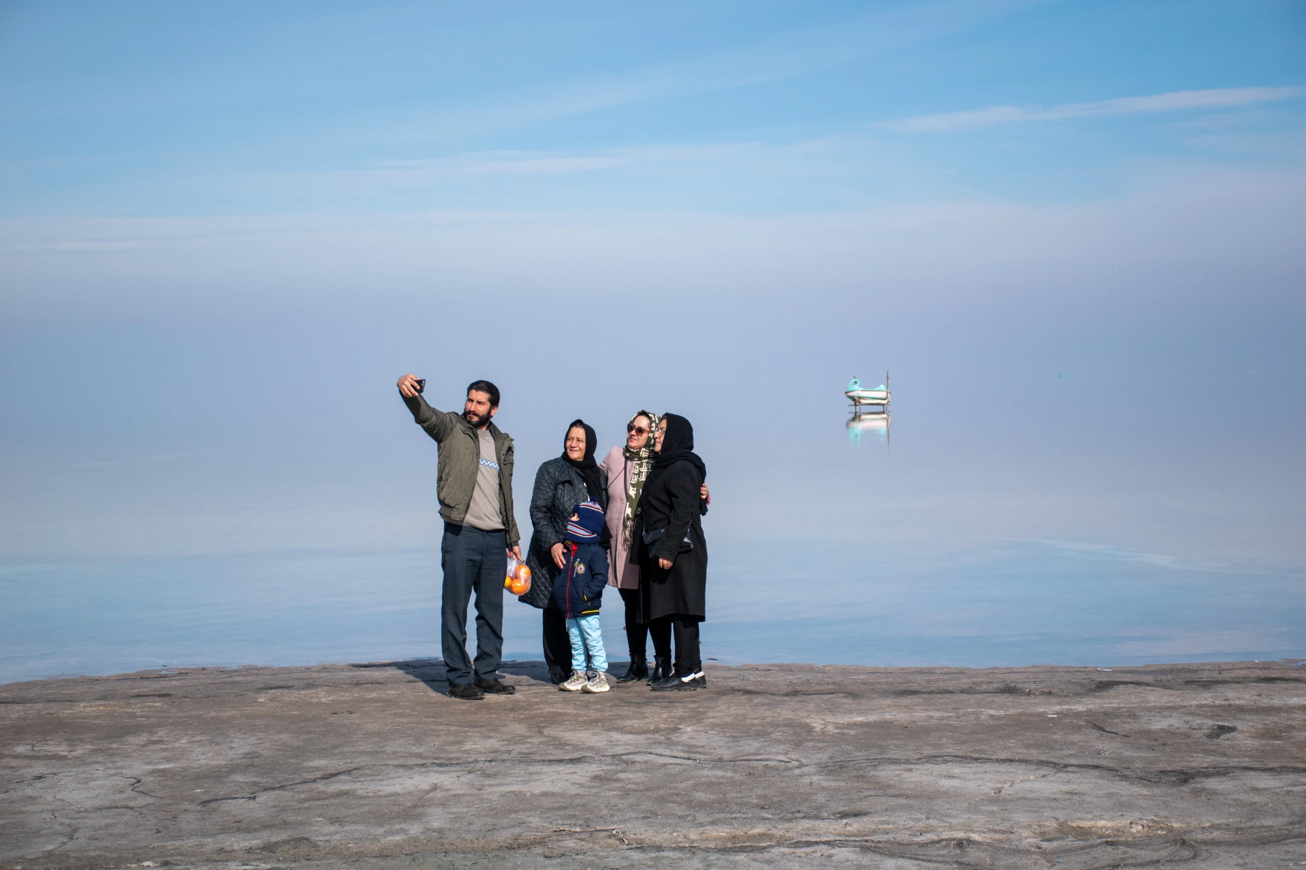A family of five stands on a rocky shore taking a selfie against the calm backdrop of Lake Urmia under a blue sky, with a small distant object reflecting on the water. Photograph by Mohammad Ataei.