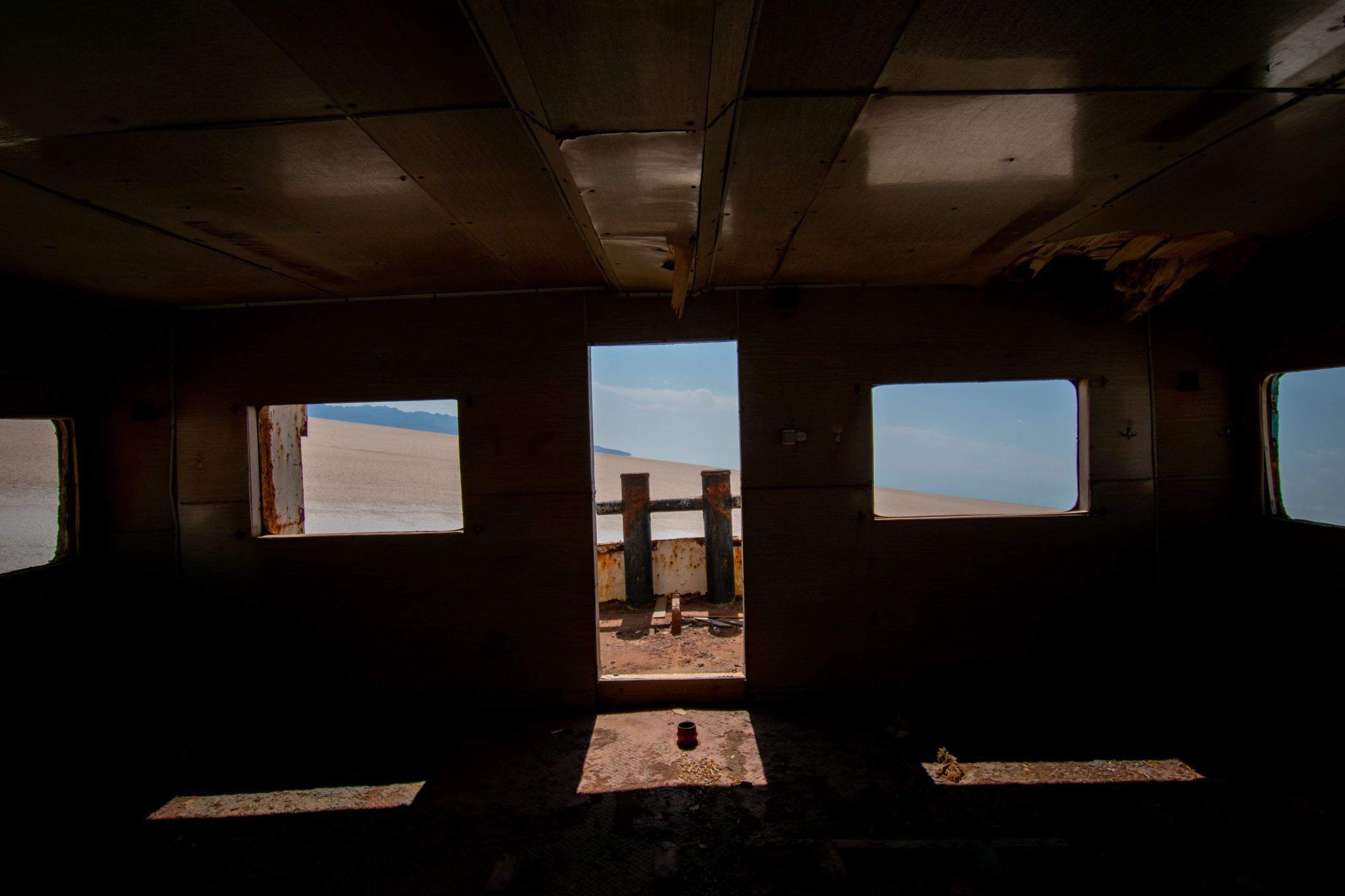Interior view from a dark, ruined cabin of a grounded ship looking out through windows and a doorway toward a bright desert or salt flat landscape under a blue sky. Photograph by Mohammad Ataei.