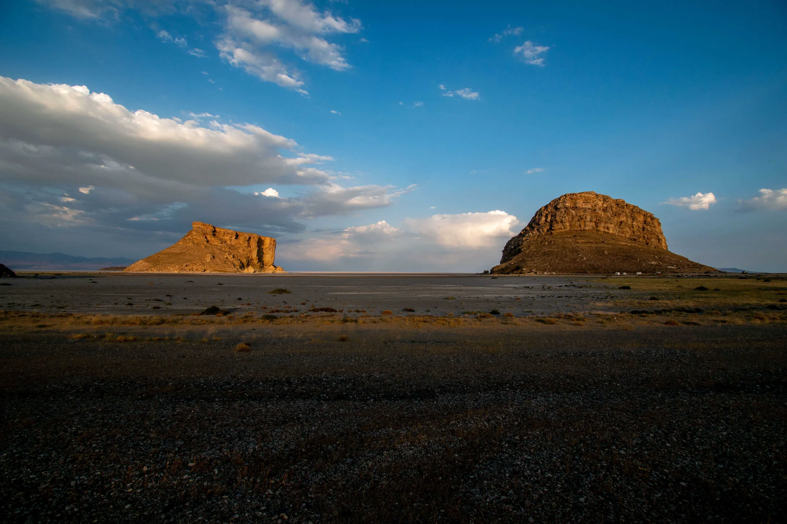 A landscape photograph showing two large rocky formations, Kazem Dashi and Qala Dashi, rising from a dry plain under a vast blue sky with scattered clouds. Photograph by Mohammad Ataei.