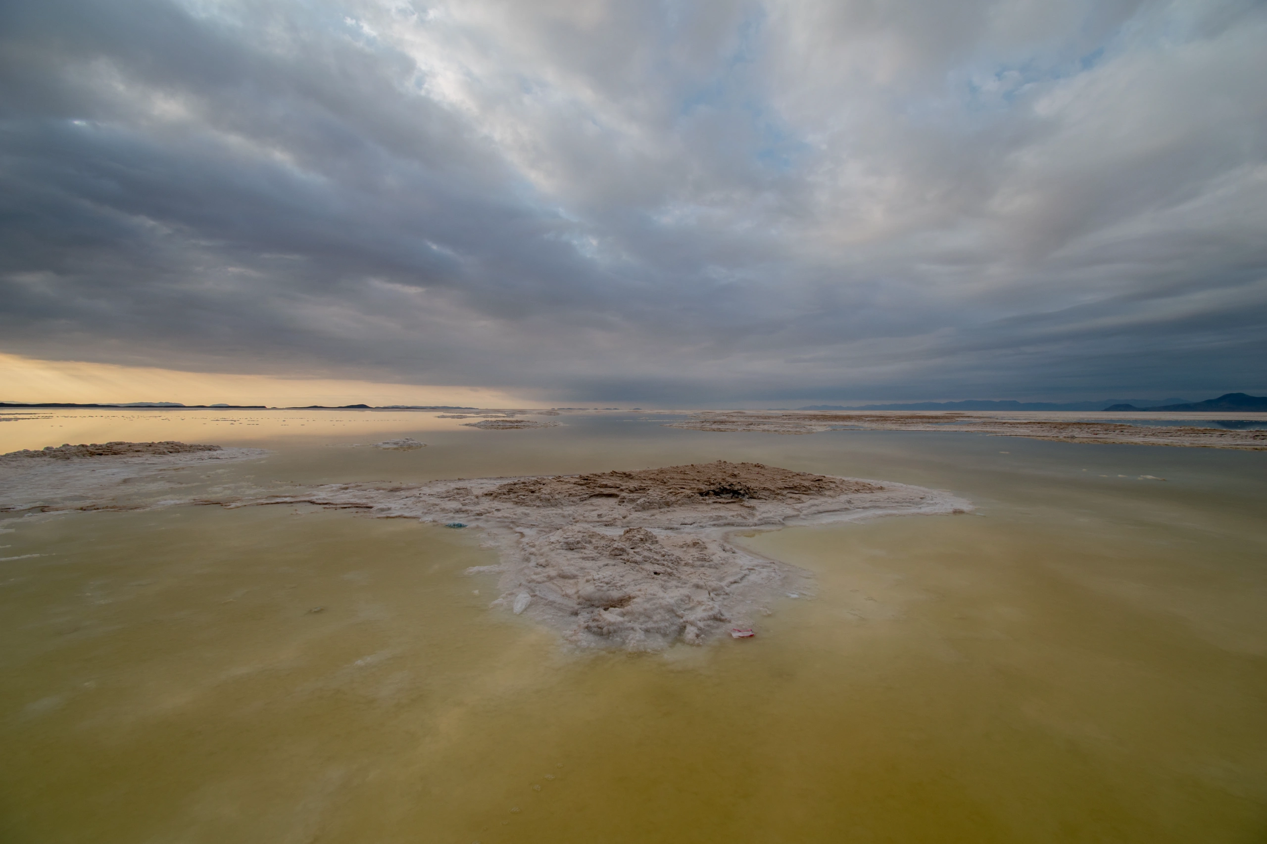 A wide-angle landscape photograph of Lake Urmia showing shallow saline waters and crystalline salt formations under a dramatic overcast sky. Photograph by Mohammad Ataei.