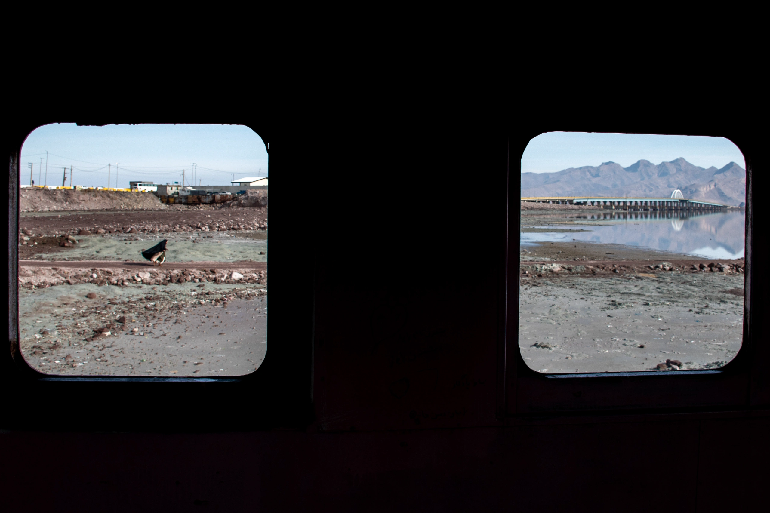 Two rectangular windows from a dark interior reveal an exterior landscape of a dry lakebed, a distant bridge, and mountains under a clear sky. Photograph by Mohammad Ataei.