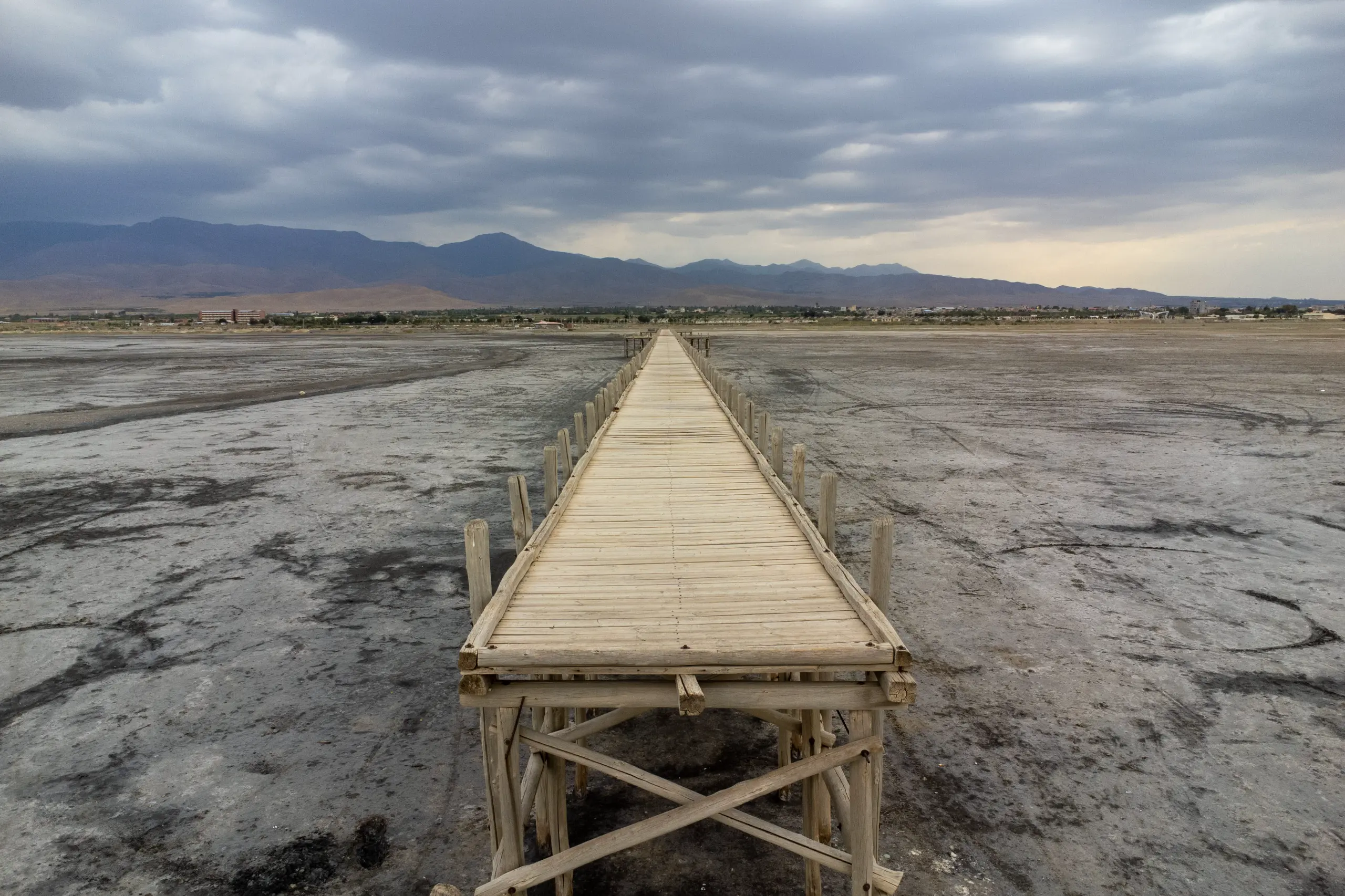 A straight wooden pier stretches across the dry, cracked bed of Lake Urmia under a cloudy sky, with distant mountains on the horizon. Photograph by Mohammad Ataei.