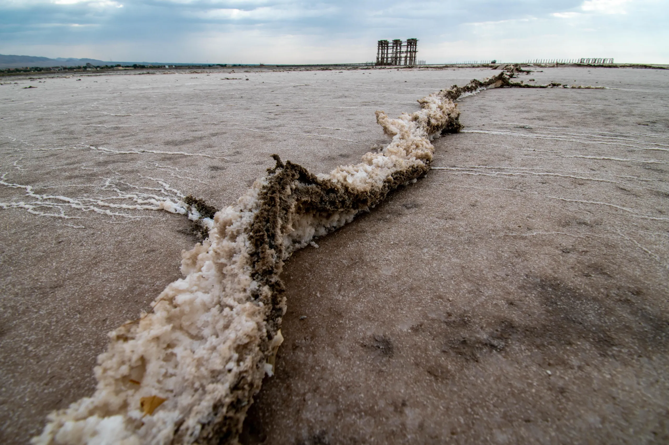 A close-up shot of a winding salt and soil ridge on the cracked, dry bed of Lake Urmia under a cloudy sky, with distant structures visible on the horizon. Photograph by Mohammad Ataei.