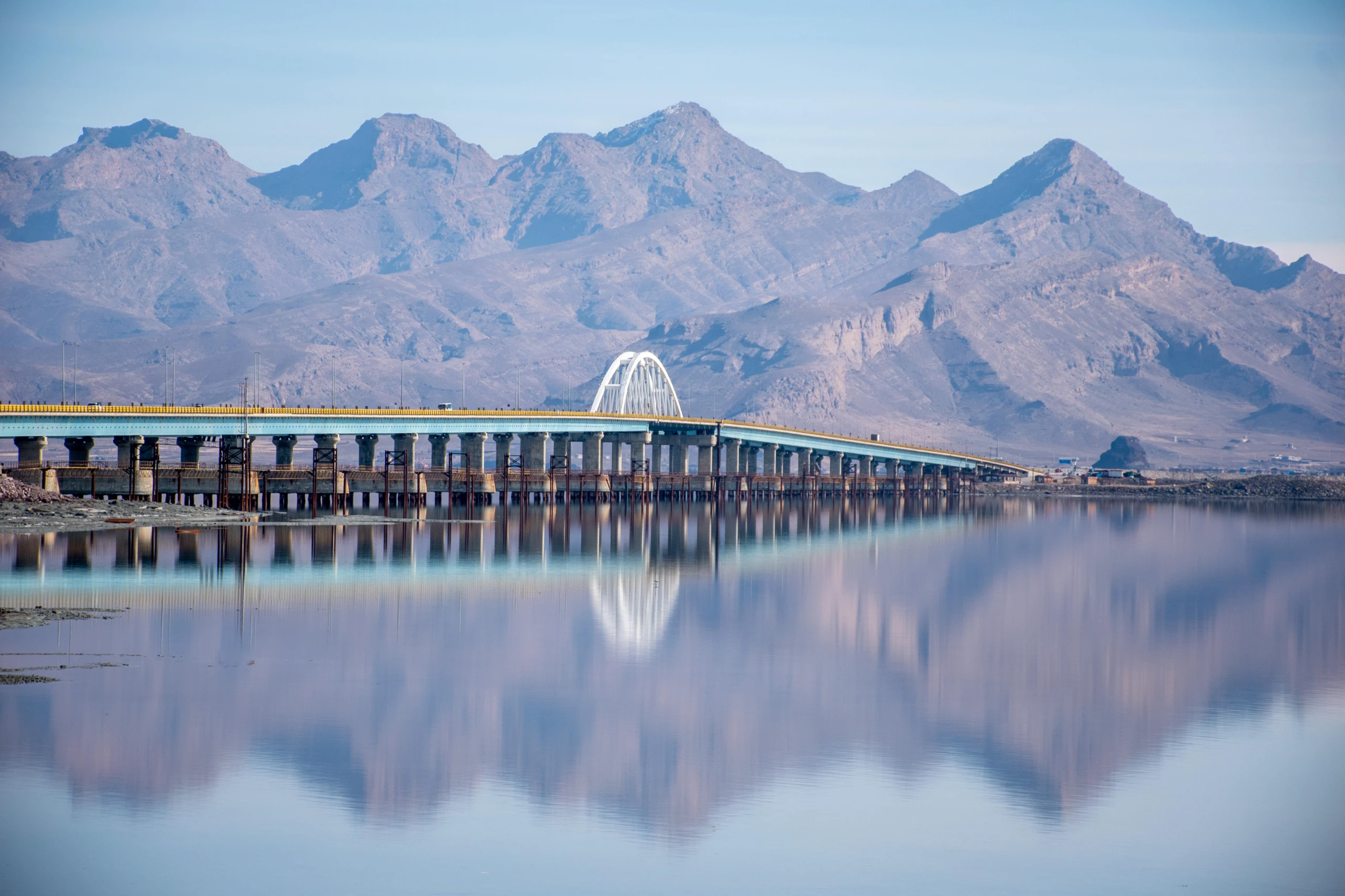 A long bridge with a white arch spans across calm water, reflecting mountains in the background under a clear sky. Photograph by Mohammad Ataei.