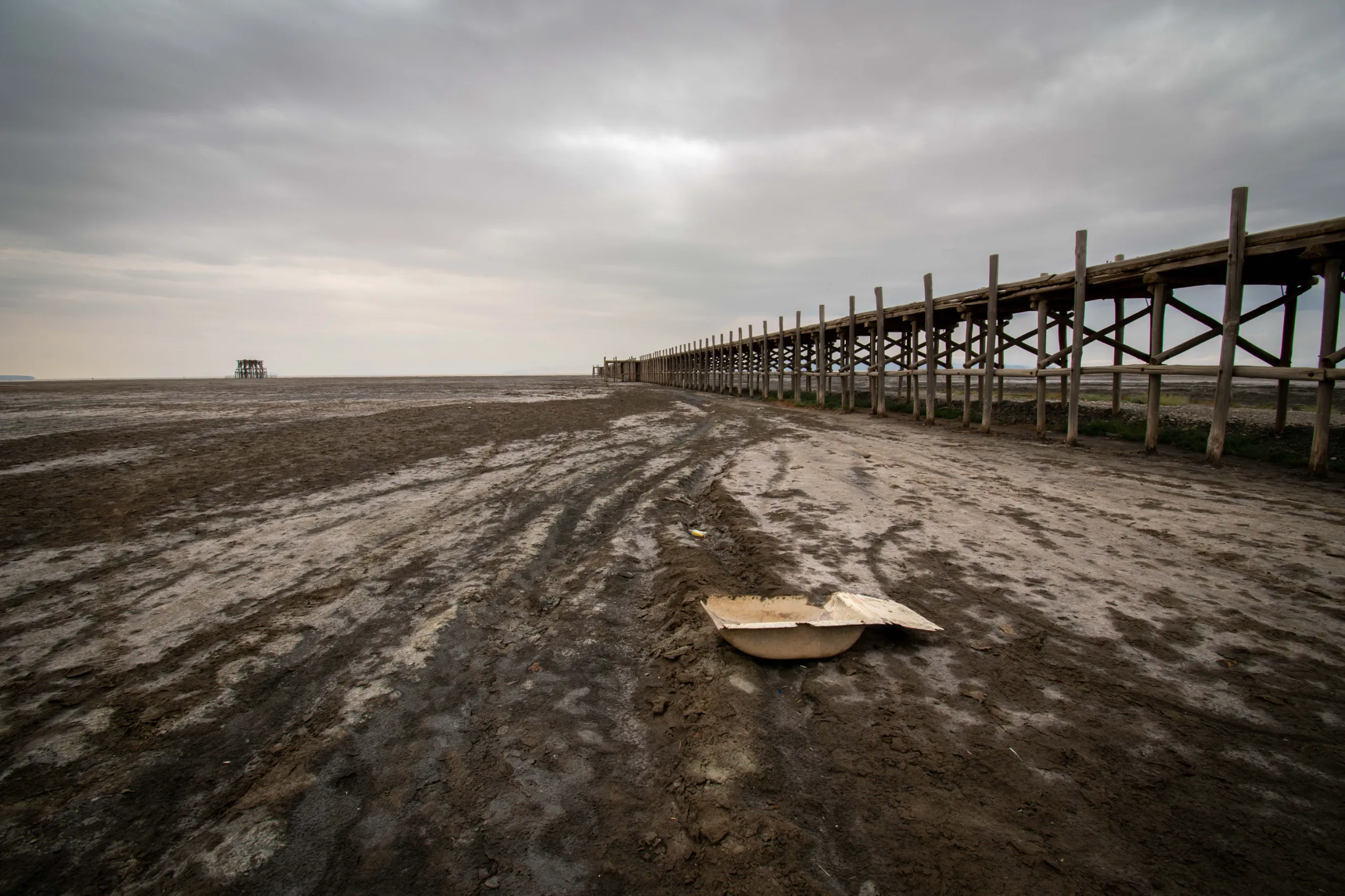 Wide angle view of the wooden Sharafkhaneh Pier extending over a dry, muddy lakebed under an overcast sky at Lake Urmia. Photograph by Mohammad Ataei.