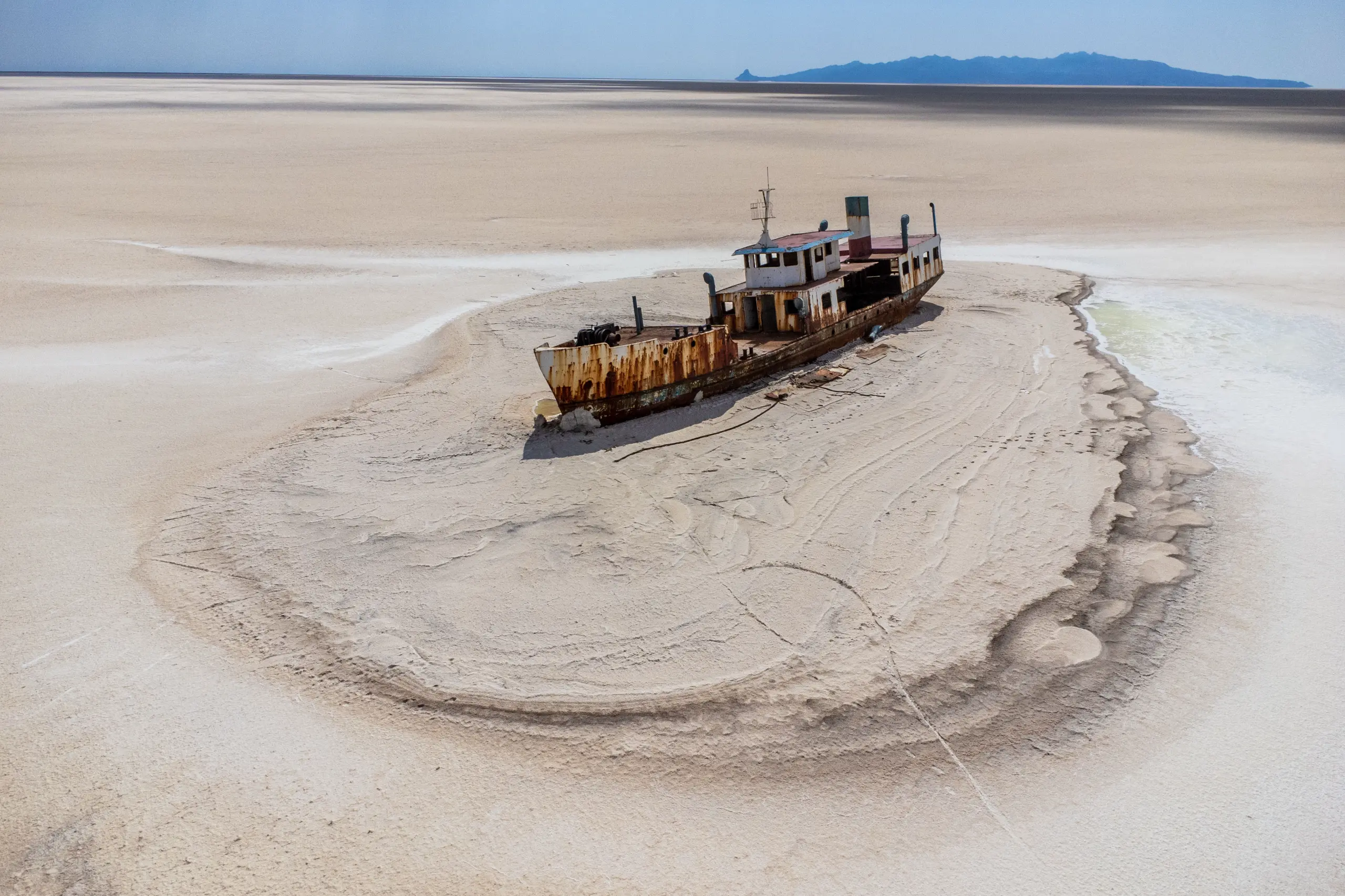 An aerial photograph shows a rusted shipwreck stranded on a dry, white salt flat surrounded by the vast expanse of Lake Urmia, with distant mountains under a clear sky. Photograph by Mohammad Ataei.