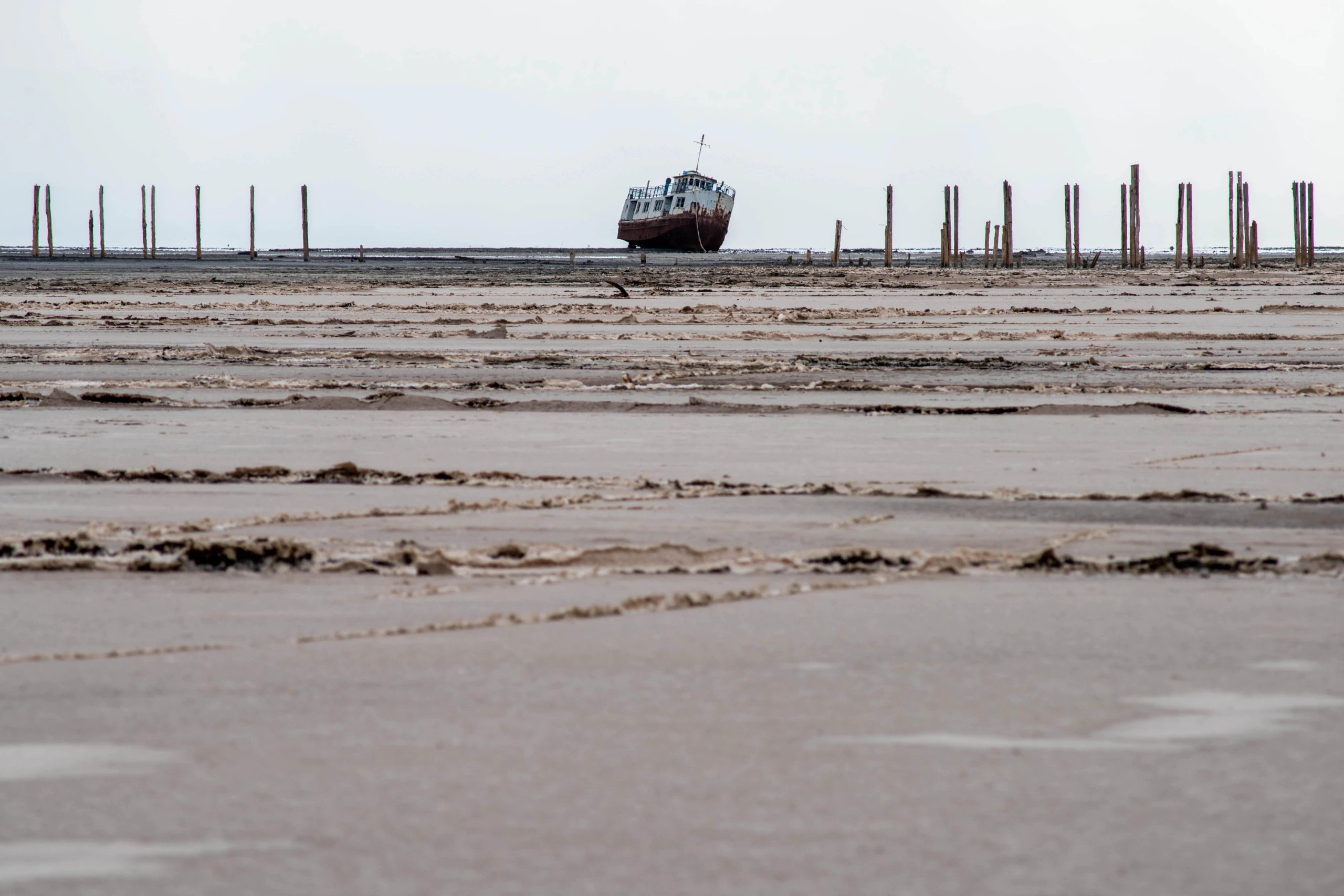 A wide-angle shot of a rusted shipwreck stranded on the barren, muddy shoreline of Lake Urmia, surrounded by wooden posts under an overcast sky. Photograph by Mohammad Ataei.