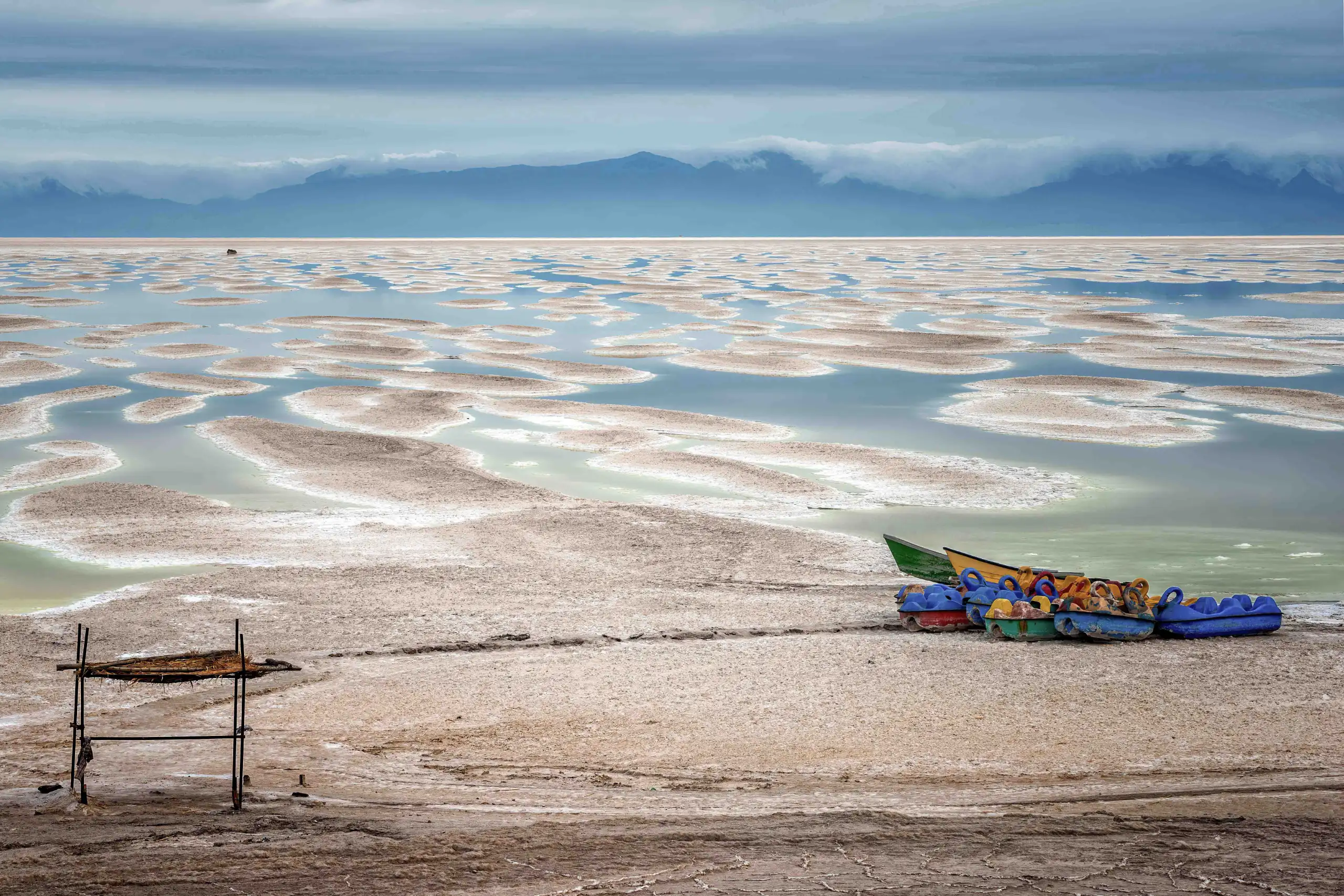 A wide landscape photograph showing the vast salt formations and shallow water of Lake Urmia under a cloudy sky, with colorful boats and a small wooden structure on the shore. Photograph by Mohammad Ataei.