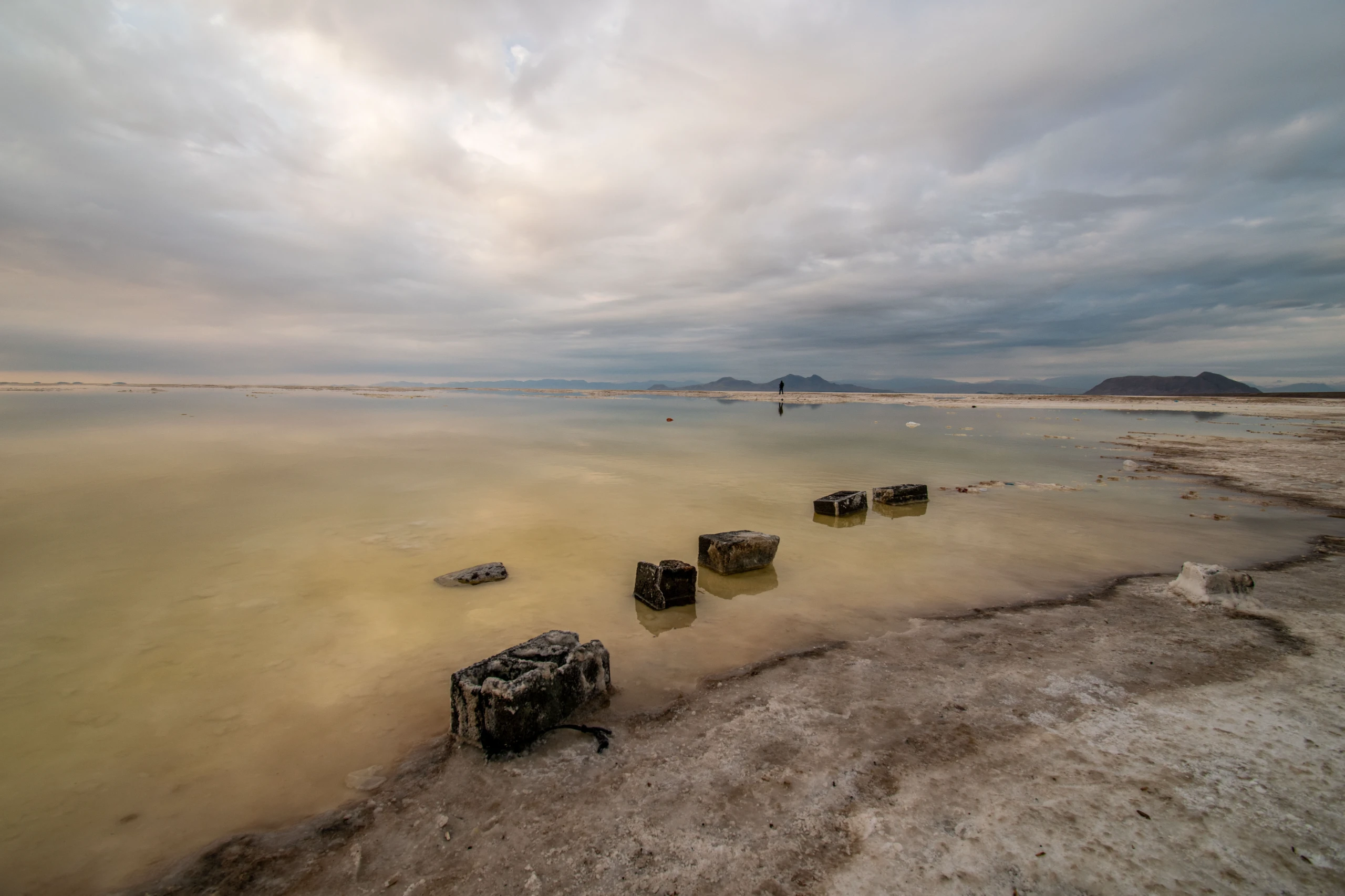Wide-angle view of the shallow shoreline and dark blocks extending into Lake Urmia under a cloudy sky. Photograph by Mohammad Ataei.