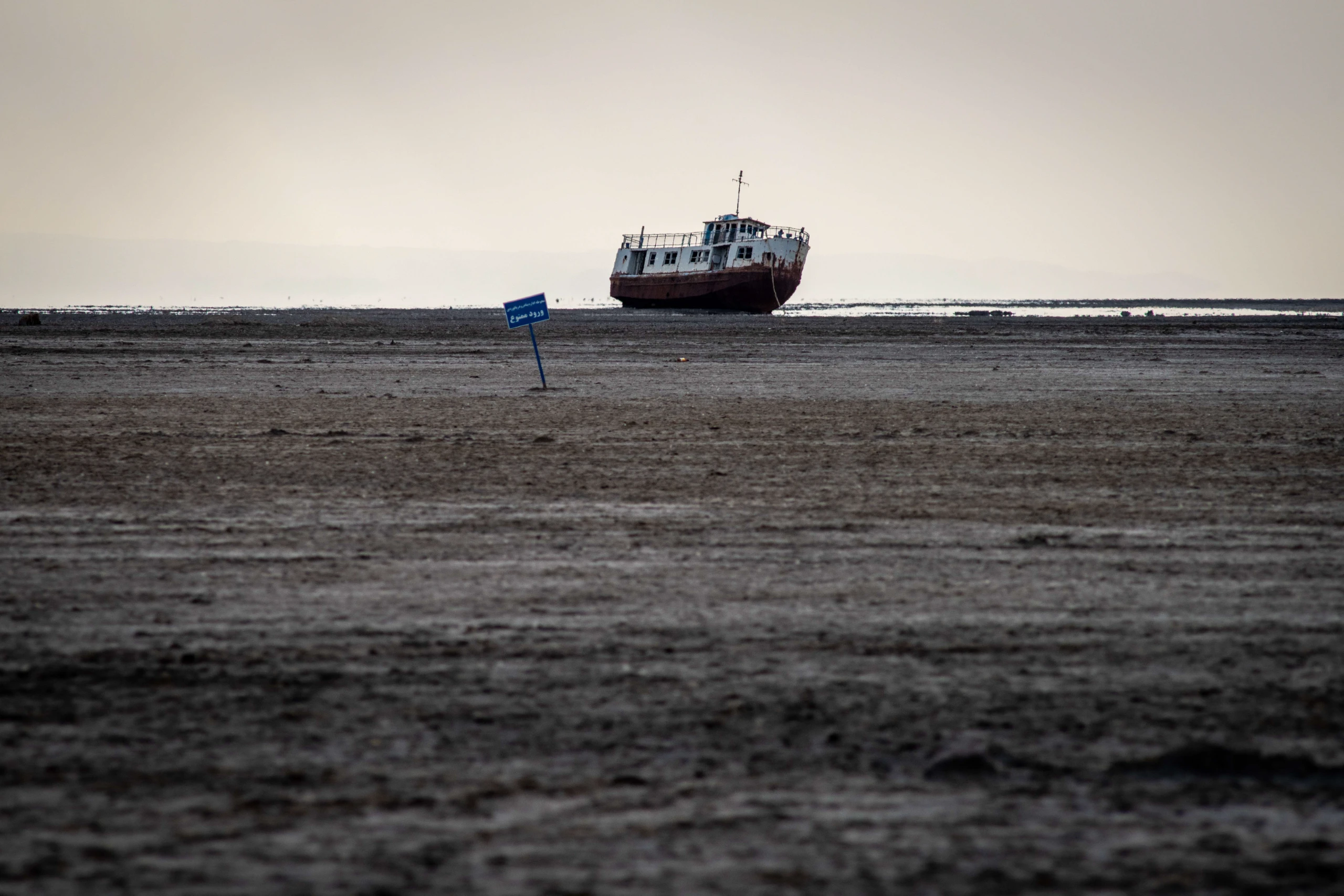 A lone abandoned ship resting on the dry, desolate bed of Lake Urmia under an overcast sky, with a small blue sign planted nearby. Photograph by Mohammad Ataei.
