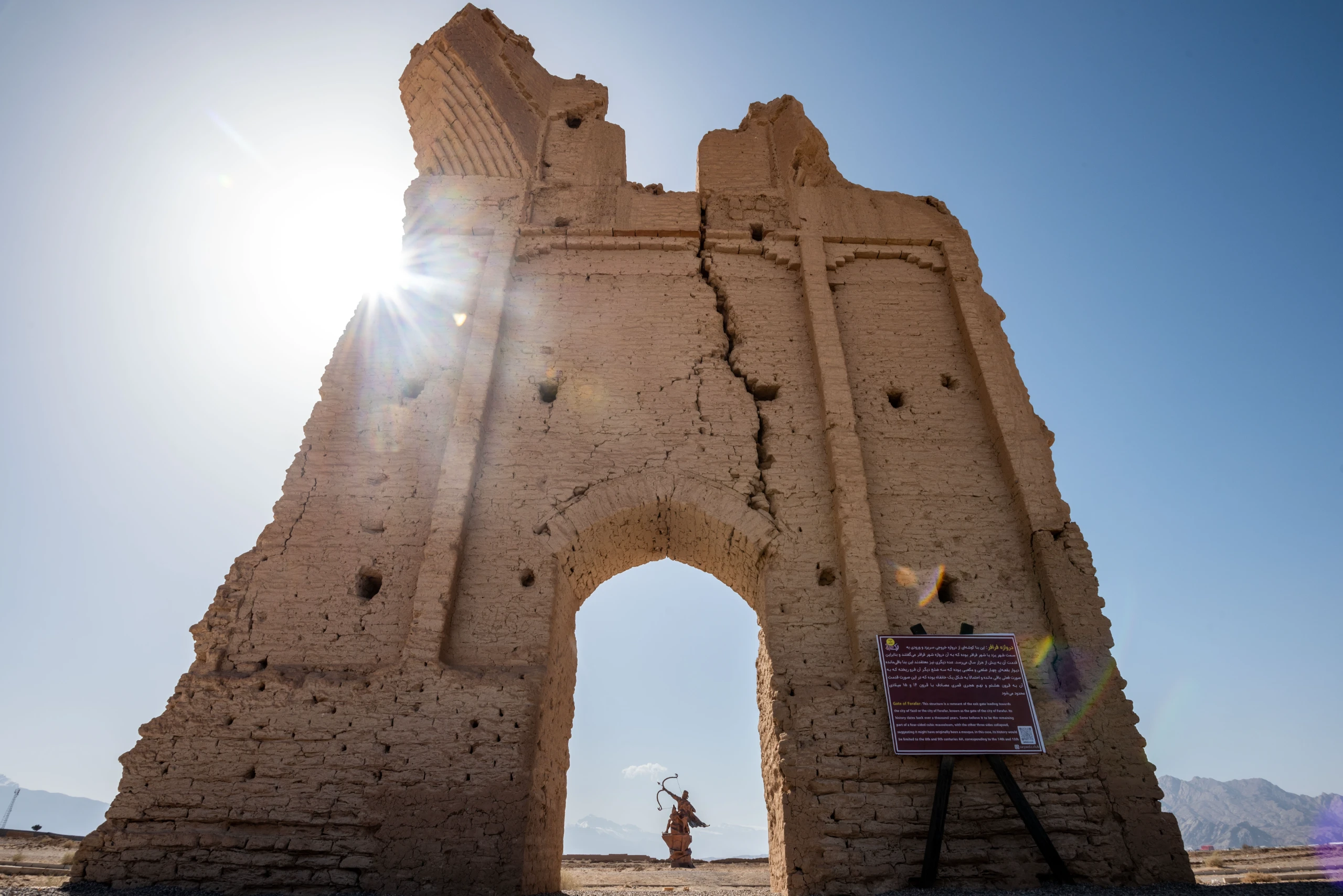 A wide shot looking up at the majestic, partially ruined Farafar Gate, an ancient mud-brick structure with a large archway, against a bright, sunny sky. A silhouette of an archer statue is visible within the arch. Photograph by Mohammad Ataei.
