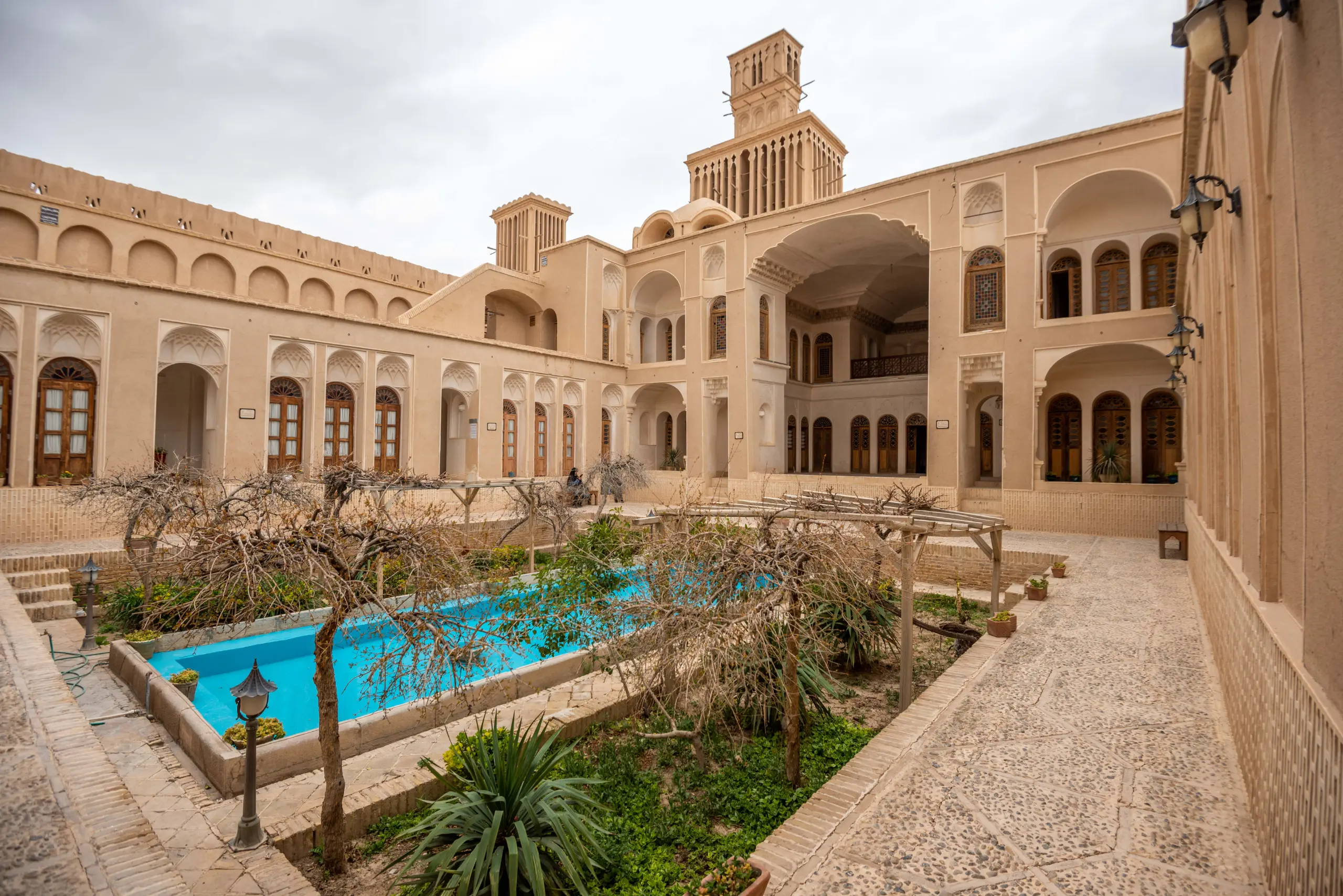 A wide shot of the courtyard of a traditional Persian adobe house with a blue central pool, surrounding gardens, and a prominent double-tiered windcatcher on the roof. Photograph by Mohammad Ataei.