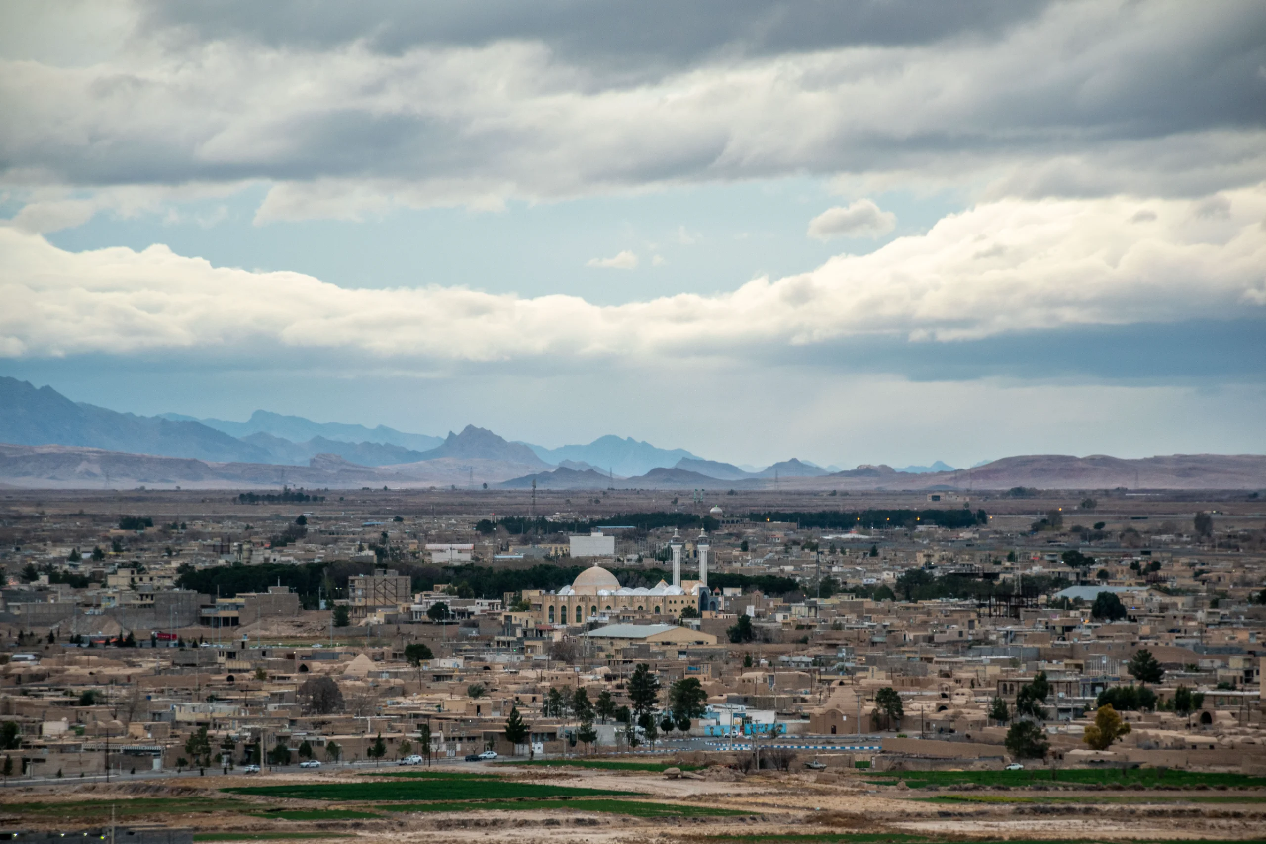 An expansive, elevated view of Abarkuh city in Iran, featuring traditional buildings, a central mosque with a dome and two minarets, and distant mountains under a cloudy sky. Photograph by Mohammad Ataei.