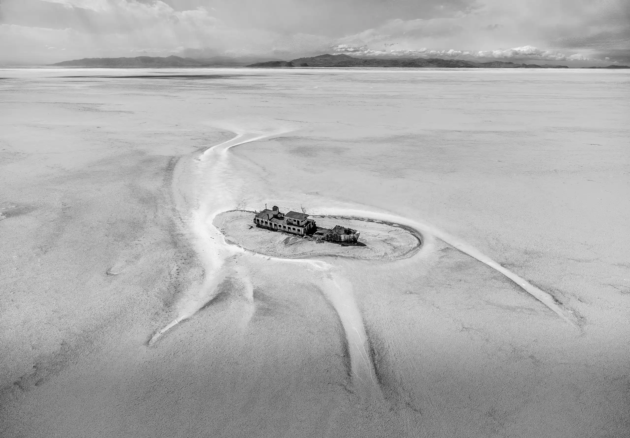An aerial drone shot features the abandoned recreational ship Artemis, red-roofed and white-sided, resting on the light brown, cracked dry bed of Lake Urmia. White winding streaks of residual water surround the vessel, stretching across the vast, flat landscape under a partly cloudy sky. Photograph by Mohammad Ataei. - Mono